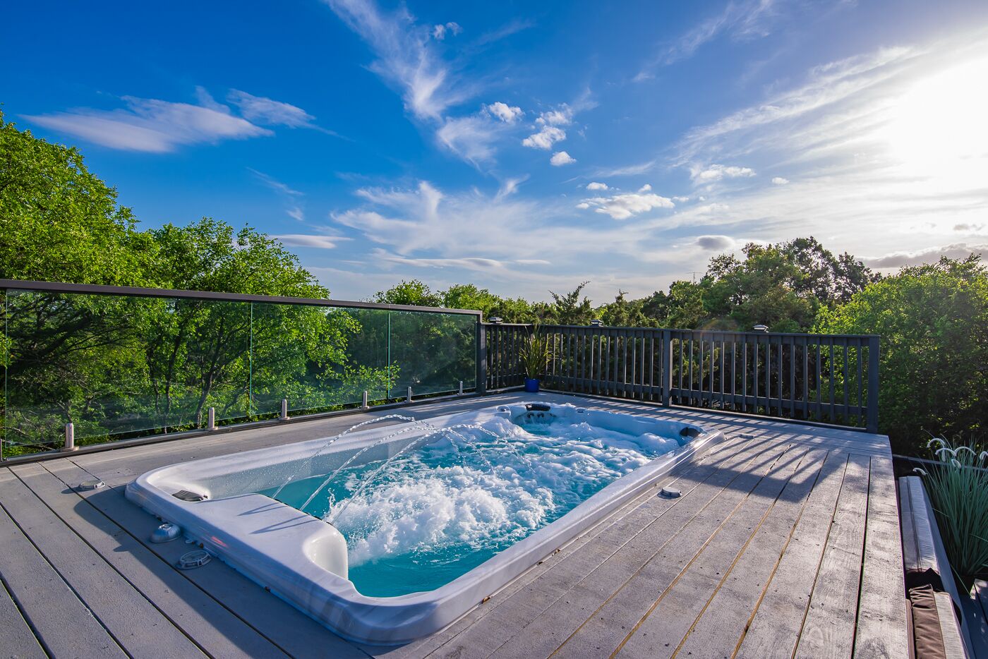 Un jacuzzi en una terraza de madera con barandilla de vidrio, rodeado de árboles.