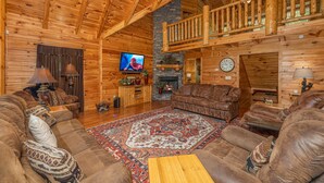 A log cabin interior with a wooden ceiling, a stone fireplace, a television, and a large rug. - Large Living Area| Game Room| Secluded| Easy Access (SEVIERVILLE)