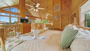 Ein Schlafzimmer einer Holzhütte mit Bett, Deckenventilator, Fernseher und Blick ins Freie.