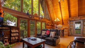 Ein gemütliches Blockhaus-Interieur mit Holzbalkendecke, Kamin, Ledersofa, Couchtisch und einer Bücherwand voller Bücher.