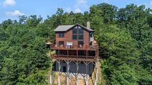 A two-story wooden cabin with a balcony, surrounded by dense forest. - Pigeon Forge*Luxury Plus*Slps 12*Incredible MTN View*Games (PIGEON FORGE)