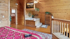 Interior de uma cabana de madeira com uma cama grande, escada de madeira, banheira de hidromassagem e ventoinha no teto.