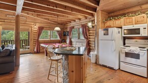 Interior de uma cabana de madeira, com cozinha equipada com frigorífico, micro-ondas e fogão. Inclui uma mesa de jantar com cadeiras e um sofá na área de estar.
