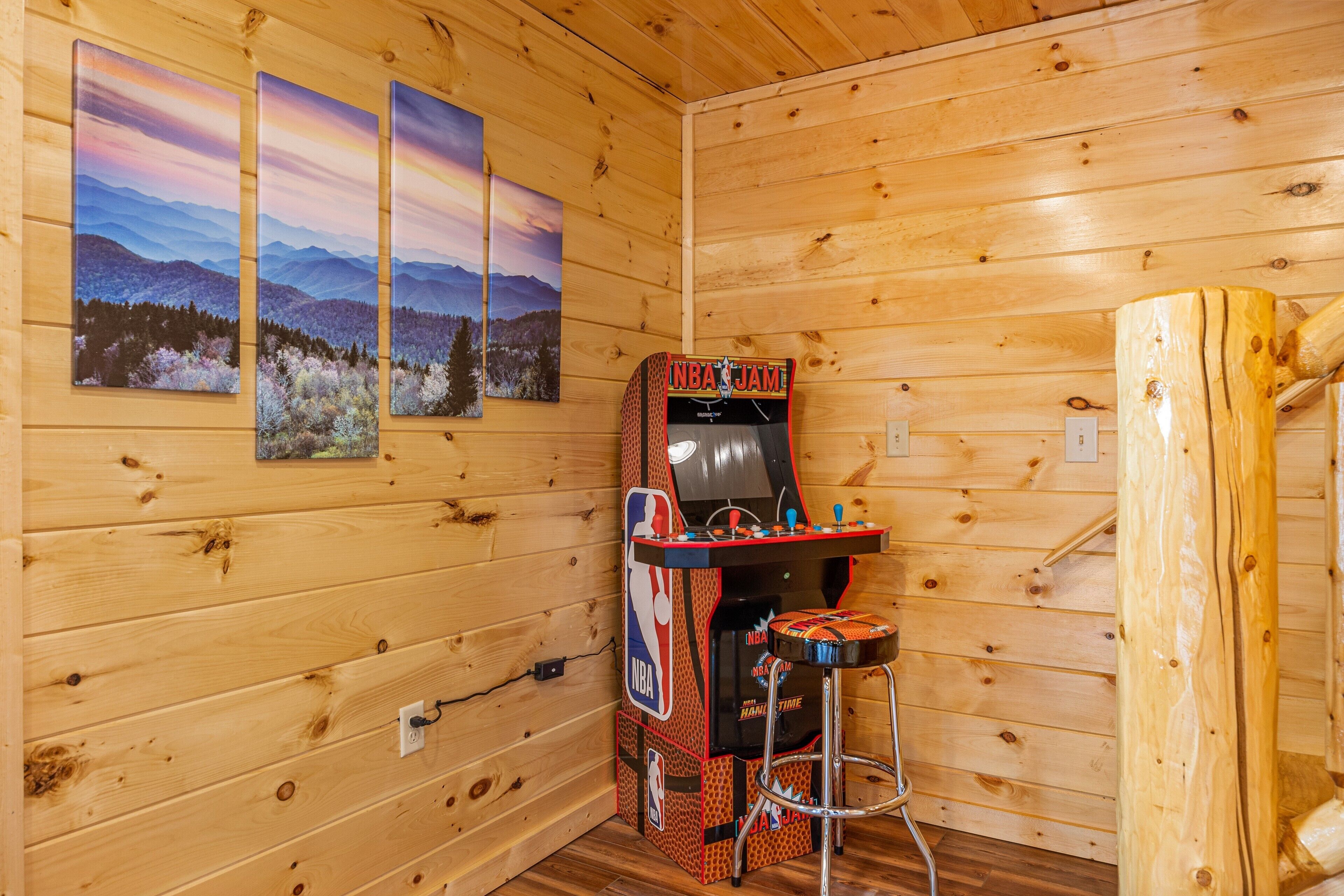Una habitación de madera con una máquina arcade de NBA JAM, un taburete de bar y tres cuadros de paisajes en la pared.