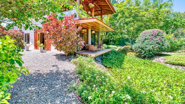 Una casa de madera con porche cubierto, rodeada de abundante vegetación y plantas en flor.