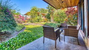 Un patio techado con mobiliario de mimbre, un jardín con diversas plantas y una vista de una casa al fondo.