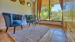 Una habitación con dos sillas azules, una mesita con un jarrón, una alfombra estampada y un ventanal con cortinas.