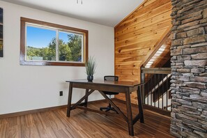 A room with a wooden desk, a potted plant, a window with a view of trees, and a stone wall.