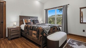 A bedroom with a large bed, a nightstand, a bench, a window with a view, and a wooden ceiling.
