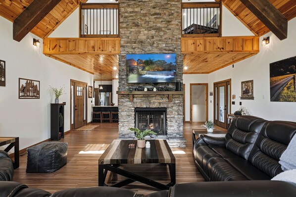 A spacious living room with a stone fireplace, wooden beams, and a large TV screen.