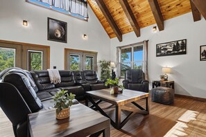 A spacious living room with a black leather sofa, a wooden coffee table, and a large window offering a view of the outdoors.