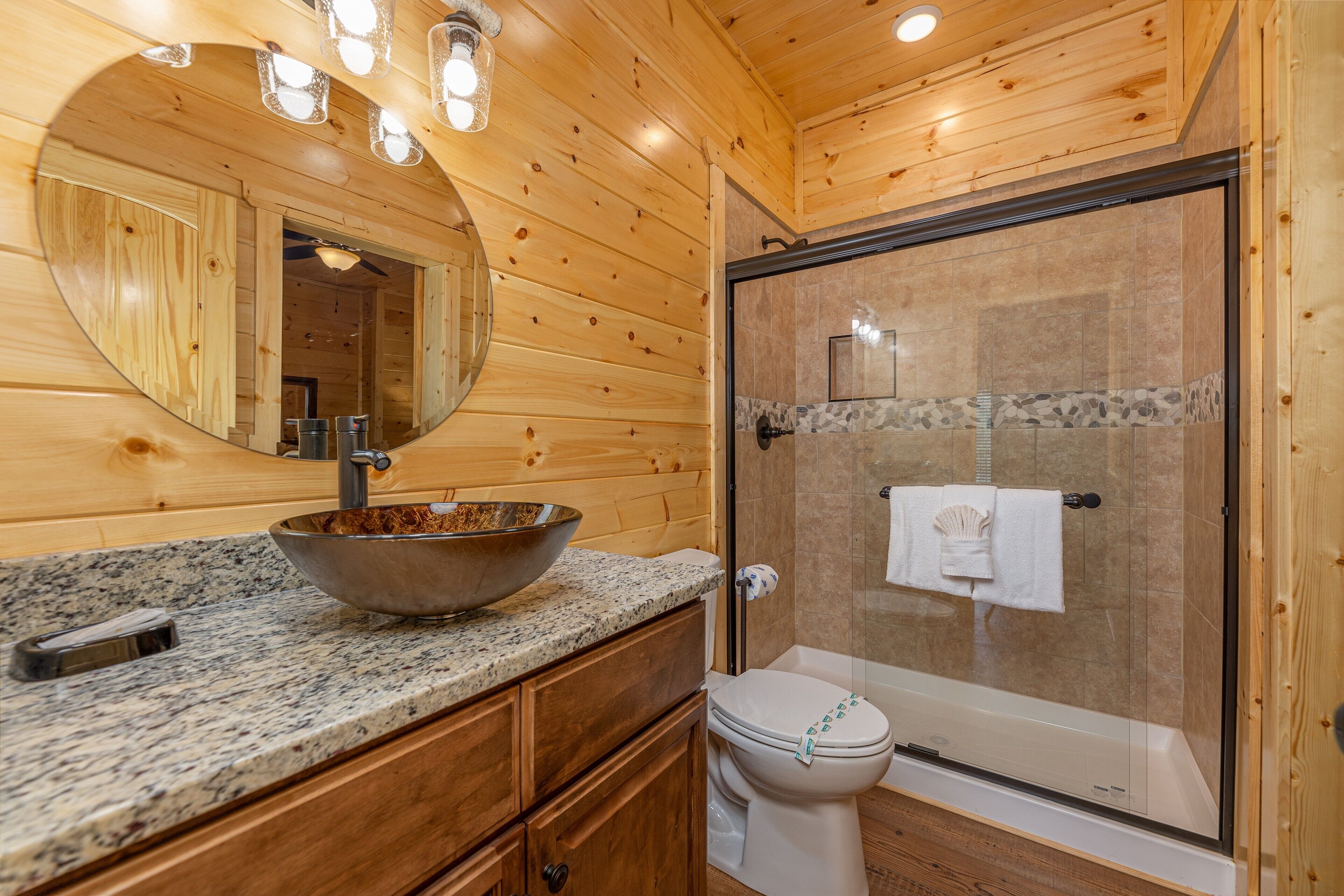 Une salle de bain rustique, avec des murs en bois, comprenant un grand miroir, un lavabo en forme de coupe, des toilettes et une douche fermée par une paroi de verre.