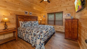 A wooden cabin bedroom with a bed, a nightstand, a lamp, a ceiling fan, a TV, and a wooden cabinet. - Mountain Views And Games Galore (SEVIERVILLE)