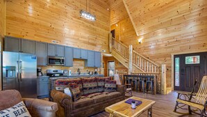 Ein gemütliches Blockhaus-Interieur mit einer Holztreppe, einer Küchenzeile, einer Couch mit gemusterten Kissen und einem Esstisch.