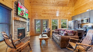 Ein gemütliches Blockhaus-Interieur mit Kamin aus Stein, Holzboden, einer Couch, Schaukelstühlen und einer Küchenzeile.