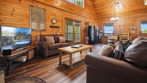 Interior de una cabaña de madera con televisor, sofá de cuero, mesa de centro de madera y un comedor.