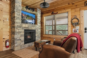 A cozy cabin interior with a stone fireplace, wooden walls, a mounted television, and a large window with blinds. - Near The Golf Corse, On The Creek (COSBY)