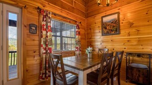 A wooden cabin interior with a dining table, chairs, a window with curtains, a welcome sign, and a framed picture on the wall. - Easy Access, Outdoor Games, Open Skies (SEVIERVILLE)