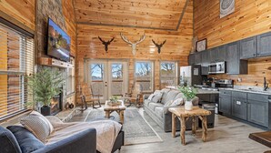 A cozy cabin interior with a fireplace, wooden furniture, a kitchen area, and deer antlers on the ceiling. - Experience Exclusivity: Unwind at Moonrise Pool Lodge Luxury Cabin (SEVIERVILLE)