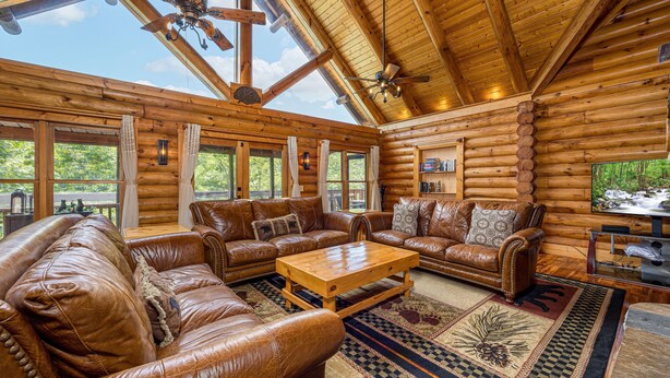 Ein Holzblockhaus-Interieur mit Ledersofas, einem Holztisch und großen Fenstern mit Blick ins Freie.