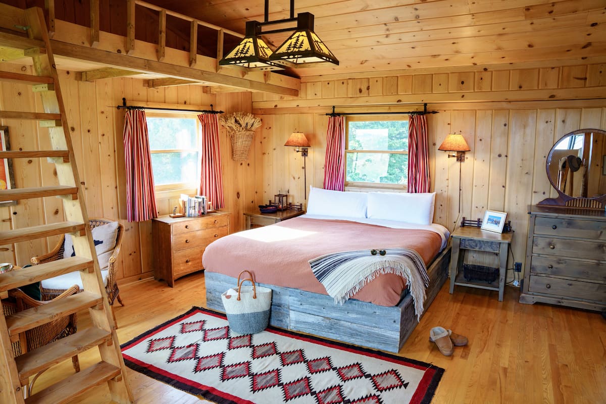 A wooden cabin bedroom with a bed, dresser, nightstand, lamp, and a mirror.
