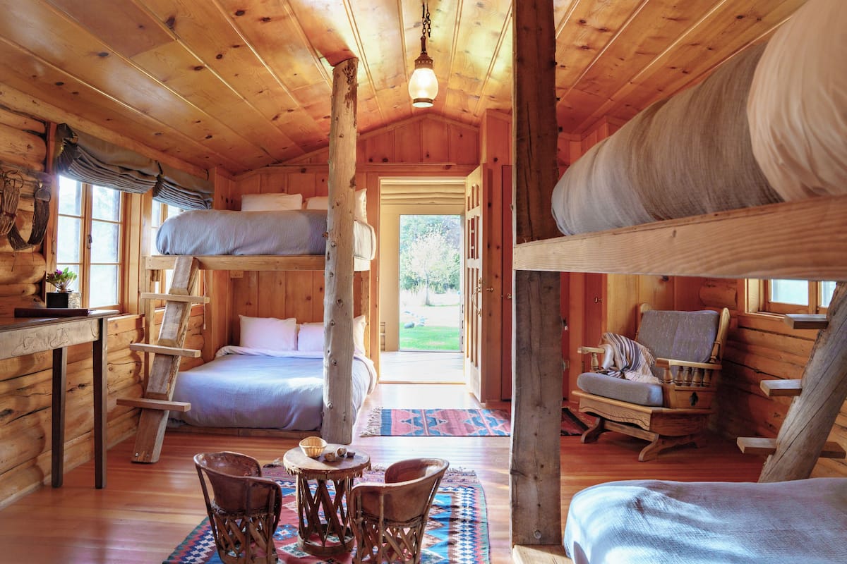 A log cabin interior with a bunk bed, a rocking chair, a dining table with chairs, and a view of the outdoors.