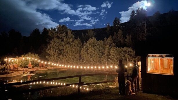 Un espacio al aire libre iluminado de noche, con un estanque, luces colgantes y una estructura de madera.