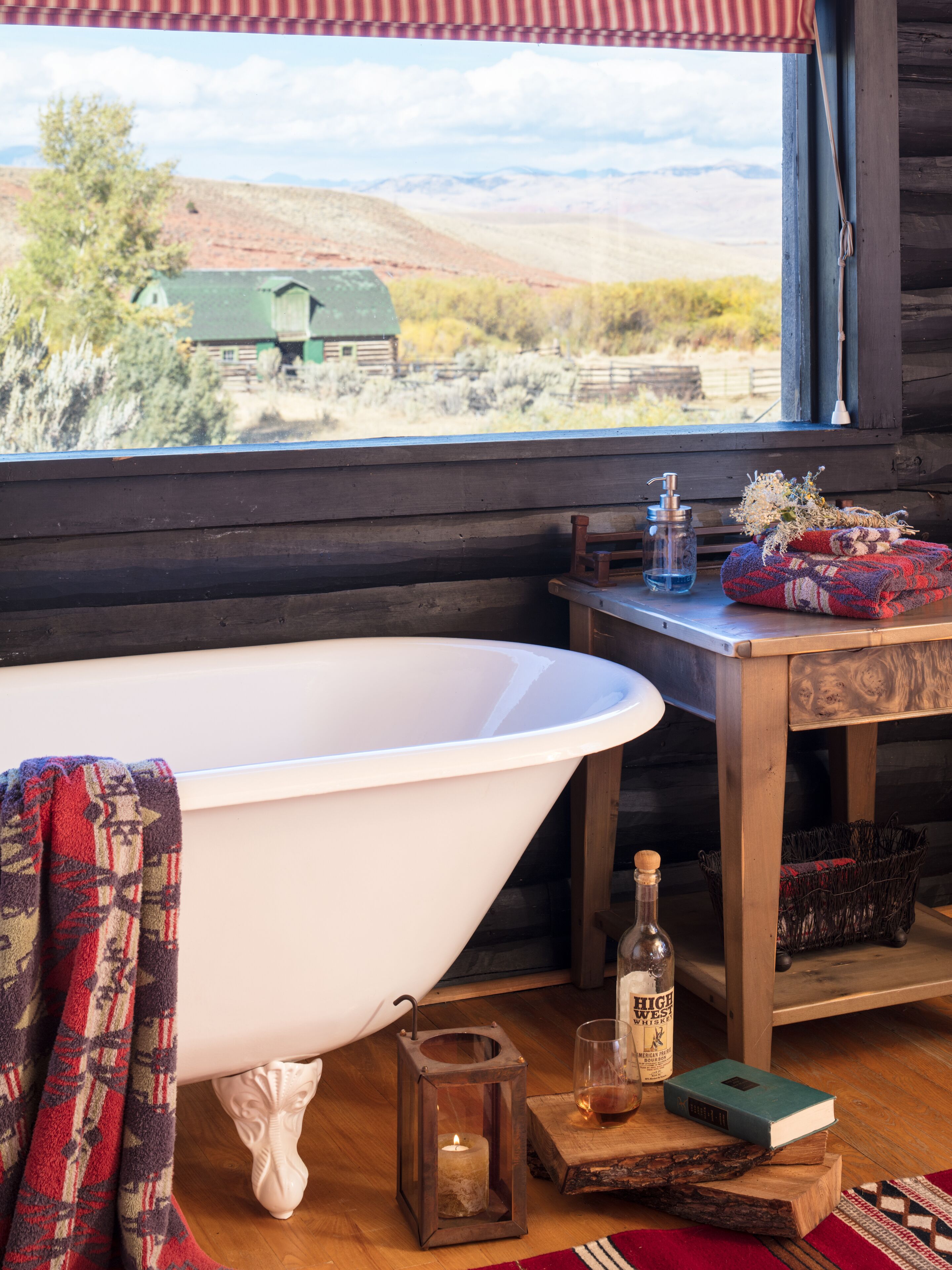 Un baño con una bañera independiente, una mesa de madera con una botella y un libro, y vista a un paisaje rural a través de una ventana.