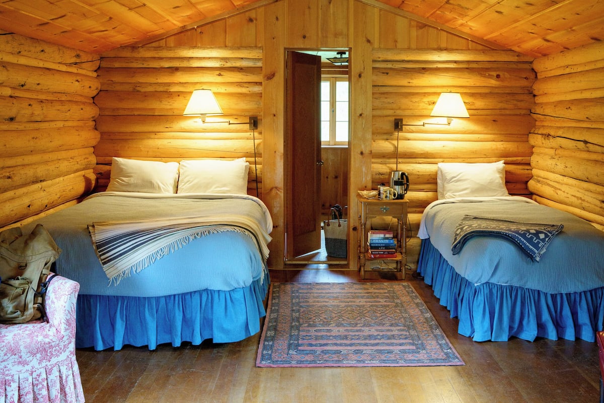 A log cabin bedroom with two beds, a nightstand, a lamp, and a rug.