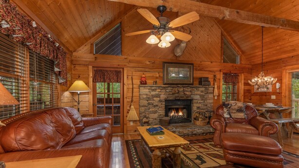 Acogedor interior de cabaña de madera con chimenea de piedra, muebles de cuero y ventilador de techo.
