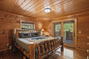 Een houten slaapkamer met een bed, een nachtkastje, een lamp en uitzicht op de natuur via een deur.