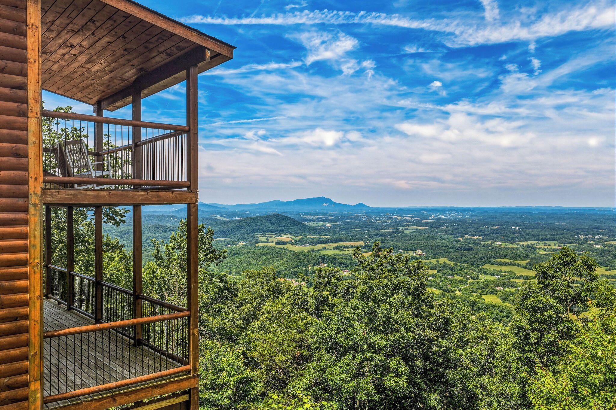 A wooden cabin with a balcony overlooking a forested valley and distant mountains.