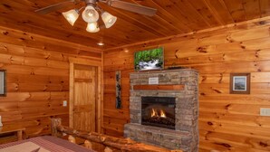 Ein Blockhaus-Interieur mit Kamin aus Stein, Holzwänden, Deckenventilator und Fernseher über dem Kamin.