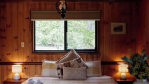A wooden-paneled room with a bed, two bedside lamps, a window with a view of trees, and a hanging light fixture.