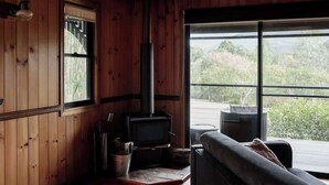 A wooden cabin interior with a wood-burning stove, a sofa, and a large window offering a view of the outdoors.