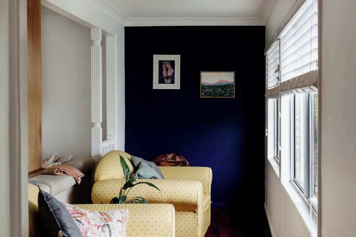 A room with a dark blue wall, a yellow patterned sofa, a white chair, and a window with blinds.