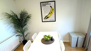 A dining area with a white table, chairs, a potted plant, a framed picture, and a fruit bowl.