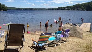 Ein Strand am Seeufer, mit badenden und spielenden Menschen, Liegestühlen und Sonnenschirmen zum Sonnenbaden.
