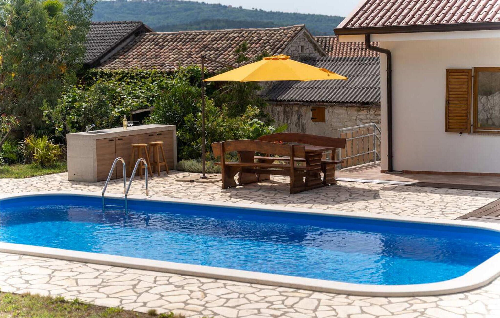 A swimming pool with a stone deck, a wooden table with chairs, a yellow umbrella, and a stone building in the background.
