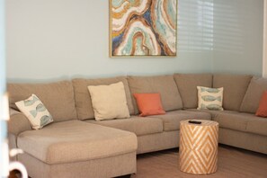 Een modulaire bank met gekleurde kussens, een rond hocker en een groot abstract schilderij aan de muur.