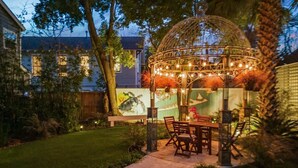 Ein Garten mit einer Terrasse, einem Pergola, Sitzgelegenheiten und einem Weg.
