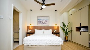 Une chambre à coucher avec un grand lit, une tête de lit en bois, des tables de chevet avec des lampes, un ventilateur de plafond et une grande plante en pot.