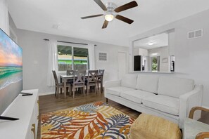 A living room with a white sofa, a dining area with a table and chairs, a ceiling fan, and a large window with a view of the outdoors. - Fenced in w/ Jacuzzi Close to Siesta Key Bridge (Sarasota)