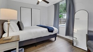 A bedroom with a bed, bedside table, lamp, ceiling fan, window with curtains, and a view of the outdoors.
