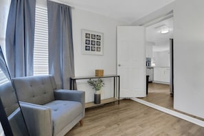 A modern living room with a grey sofa, a small table with a vase, and a door leading to a kitchen.