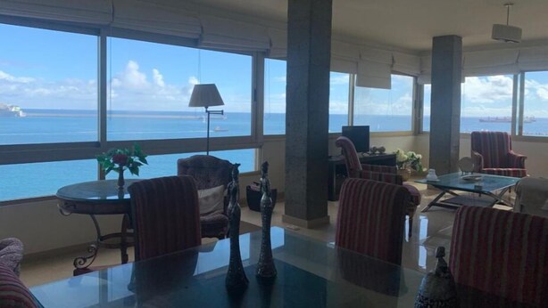 A spacious living room with large windows offering a view of the sea and ships. There are several chairs and a table with decorative items. - Azul y mar (Las Palmas de Gran Canaria)