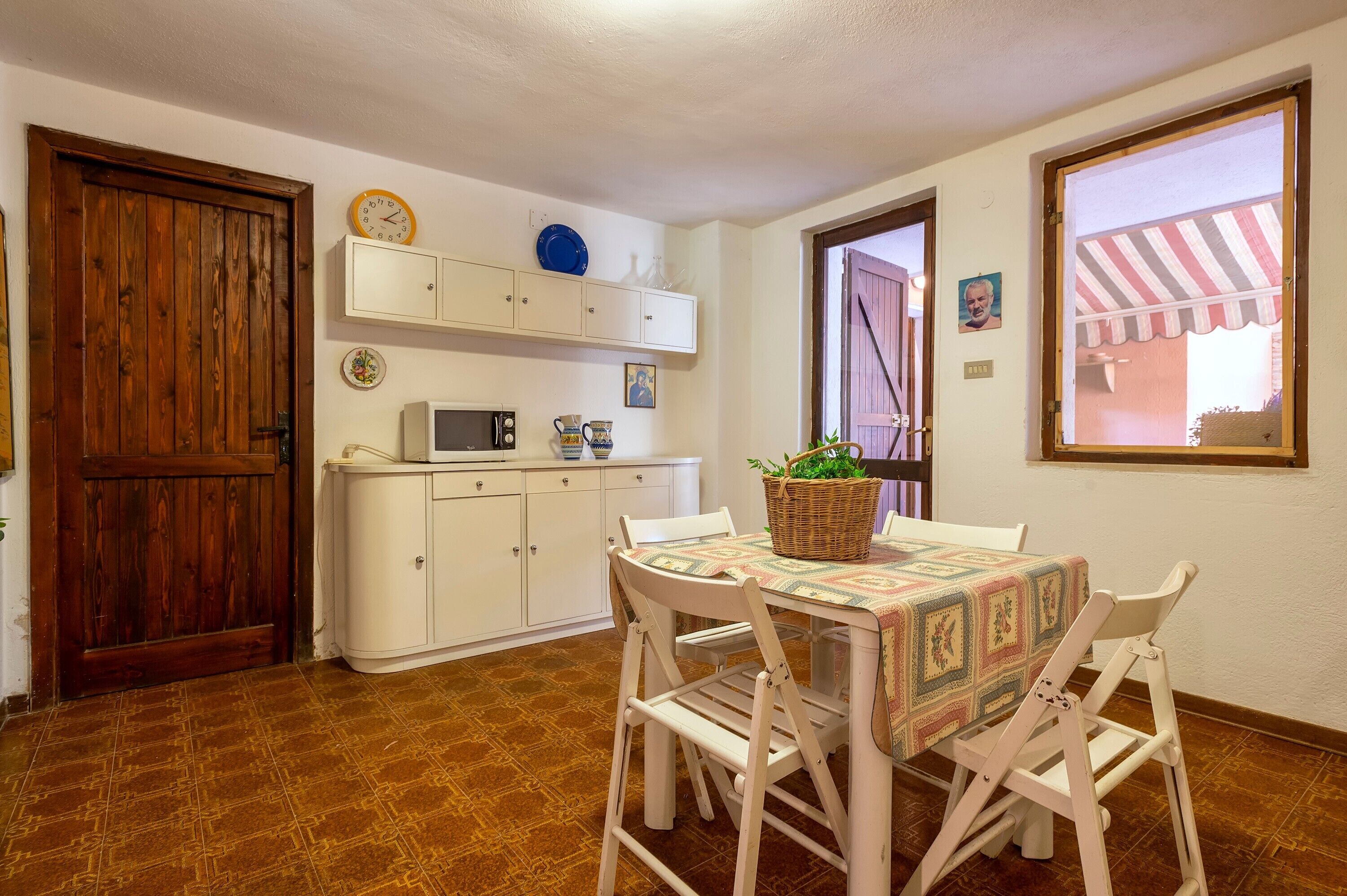 Cocina con armarios blancos, microondas y una mesa con sillas. Se observa una puerta de madera, una ventana con toldo a rayas y un reloj en la pared.