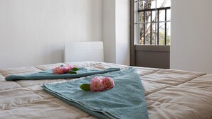 Ein Bett mit einer türkisfarbenen Decke und rosa Blumen, ein weißer Stuhl und ein Fenster mit Blick auf Bäume.