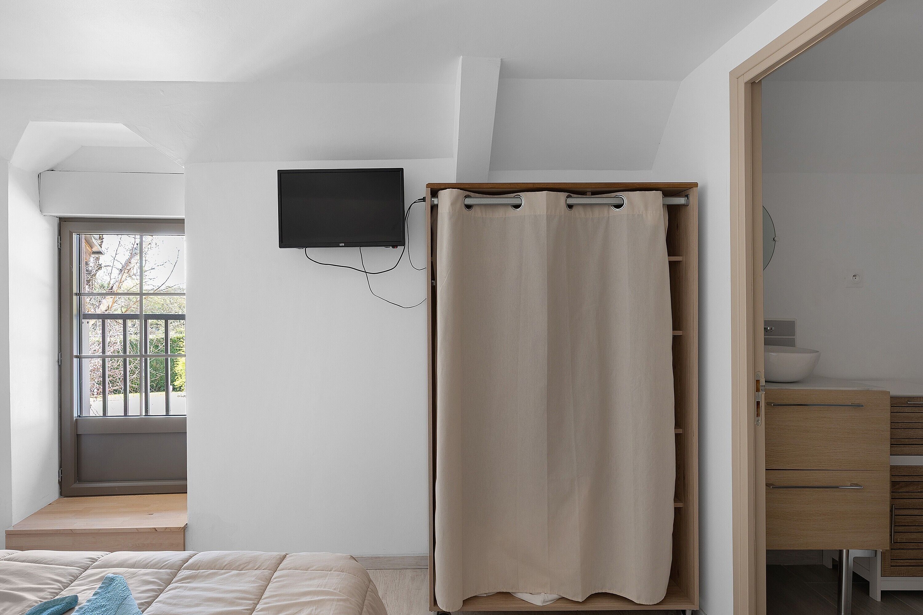 A bedroom with a bed, a TV mounted on the wall, a window, and a wooden wardrobe.