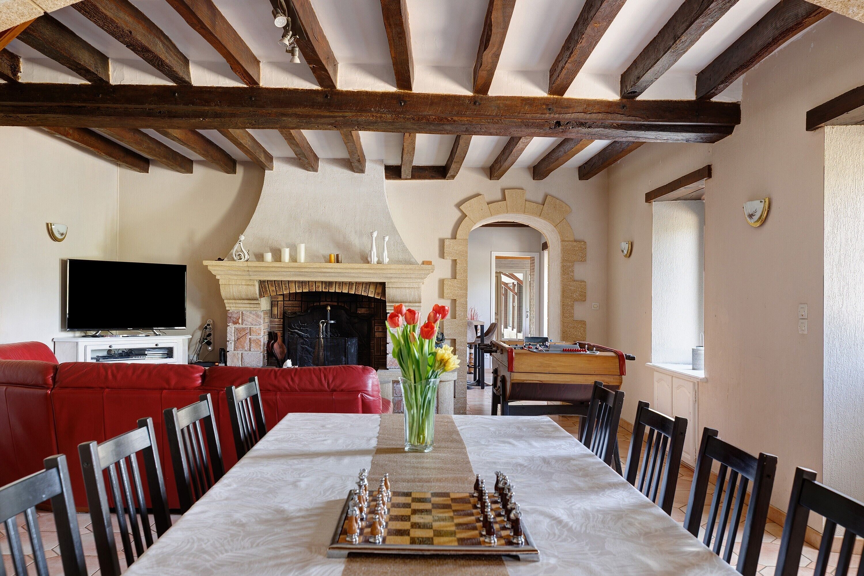 A dining area with a chessboard, a fireplace, and a television in a room with exposed wooden beams and a red sofa.